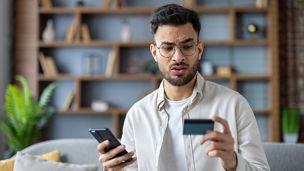 Close-up photo of a worried young Indian man sitting on the couch at home and looking frustrated at his credit card and phone.